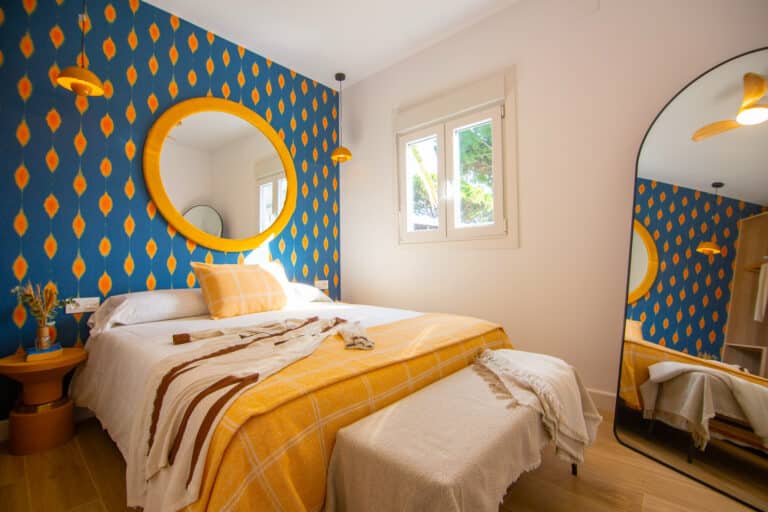 Habitación moderna con pared azul estampada, cama doble, espejo circular amarillo y ventana