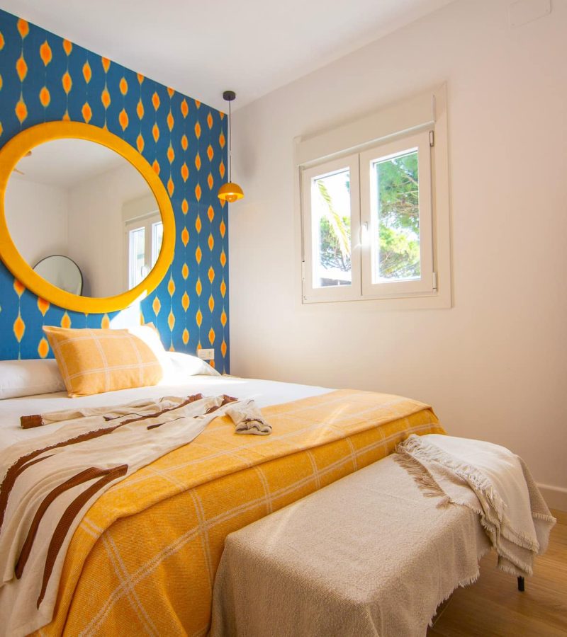 Habitación moderna con pared azul estampada, cama doble, espejo circular amarillo y ventana